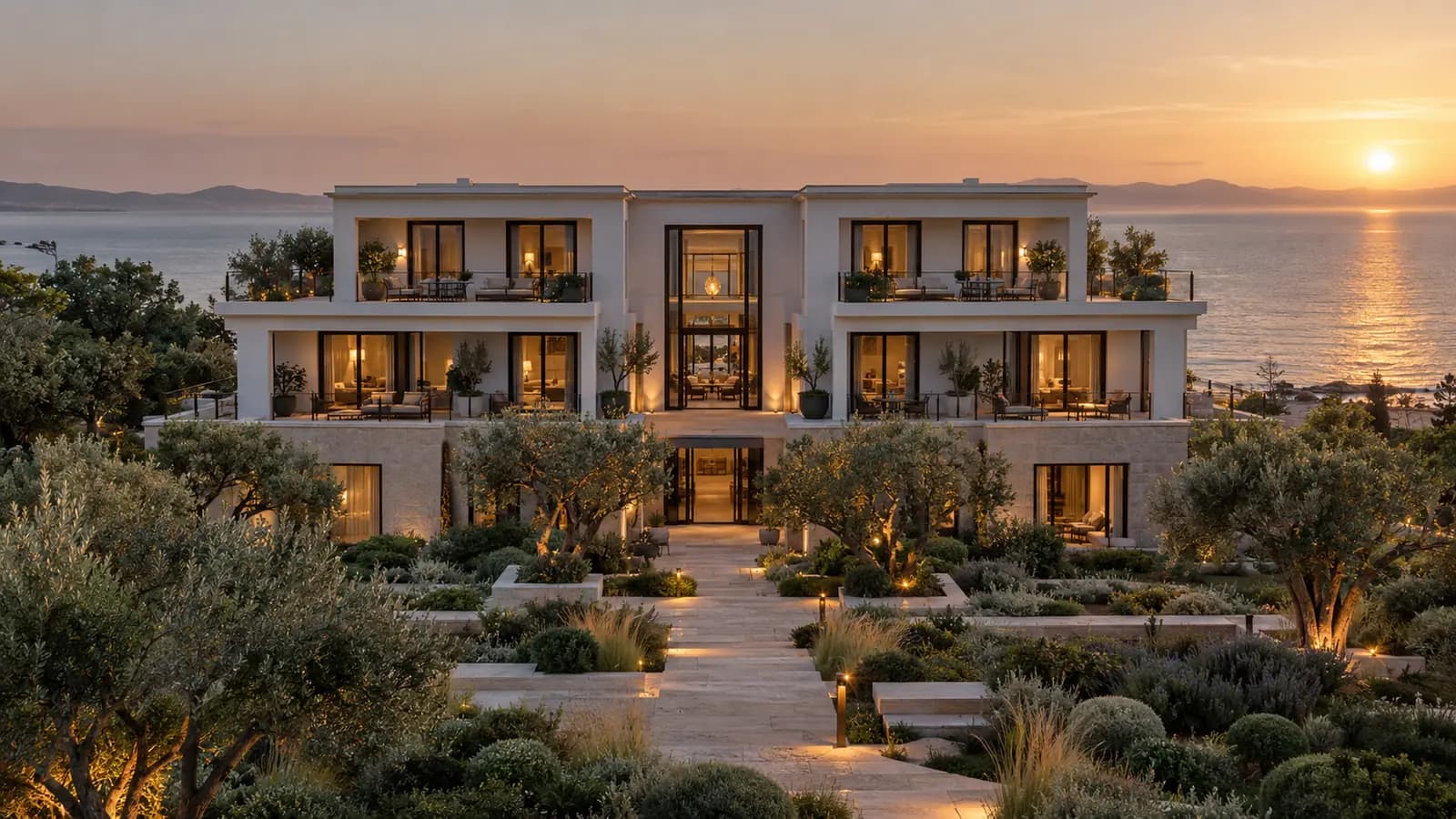 A low white hotel above the sea at sunset, olive trees and lit paths in front.